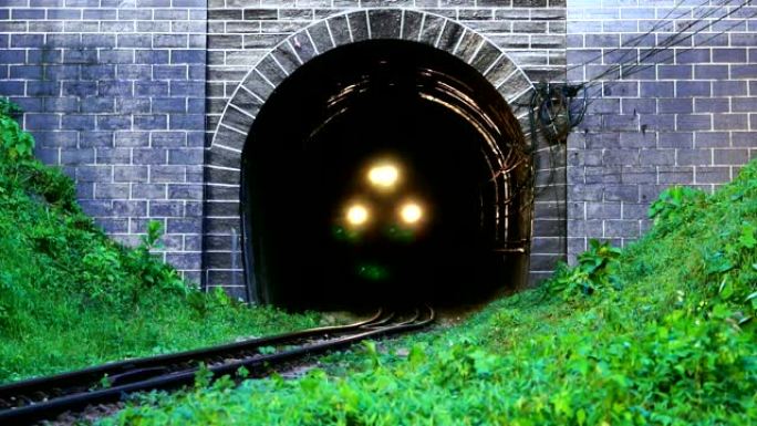 慢动作火车在吹风的情况下打开了从旧隧道行驶的前灯。古铁路隧道前山脚的绿草。