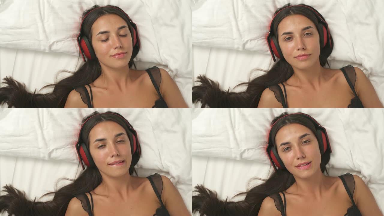 一个性感的女人在耳机躺在白色毯子上听音乐和性感地咬她的嘴唇的俯视图。