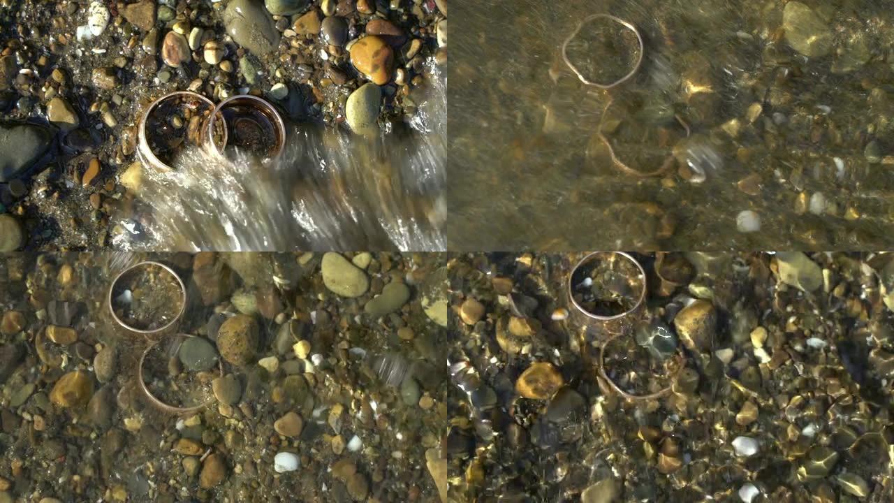 两枚金色结婚戒指躺在沙滩上的石头上，照耀着阳光，特写宏观。戒指上的光输血。海浪冲向他们，离婚观念，人
