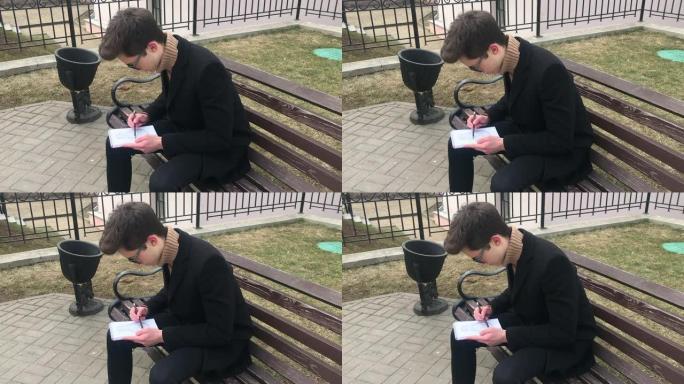 一个年轻人坐在长凳上，在笔记本上素描。春天，天气凉爽，那家伙穿着一件黑色外套。复古风格的舒适庭院。