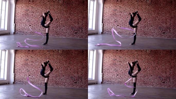 艺术体操运动员，穿着黑色紧身衣的小女孩，用丝带做杂技动作-摆着伸腿的姿势，挥舞着丝带。砖墙背景。慢动