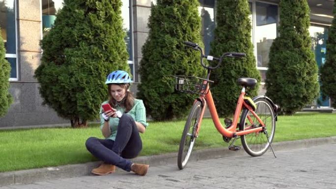 交通自行车的主题生态模式。美丽的年轻白人妇女戴着蓝色头盔和长发，站在城市公园里一辆带篮子的橙色租赁自