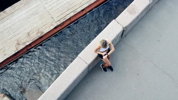 金发碧眼的运动年轻女子在慢跑并检查手腕上的健身追踪器后休息时靠在河边的路堤栏杆上的正上方视图