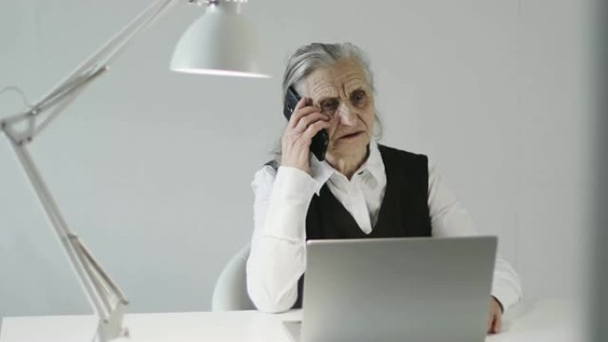 一位头发花白，皱纹很深的严肃老妇人正在办公室里用手机聊天。