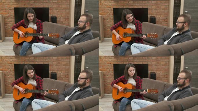 一对年轻夫妇玩得很开心，女孩坐在沙发上弹着原声吉他，而她的男朋友听着，喜欢它