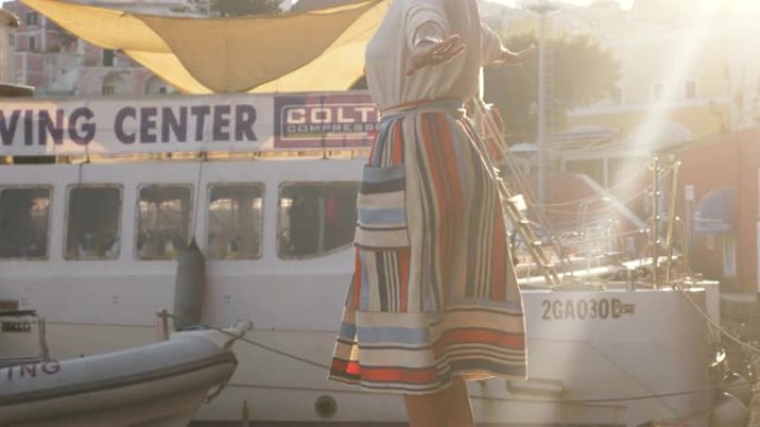 年轻女子在意大利蓬扎岛港口边的一艘船上行走在木制t台行人天桥上。时尚彩色连衣裙裙子和帽子。