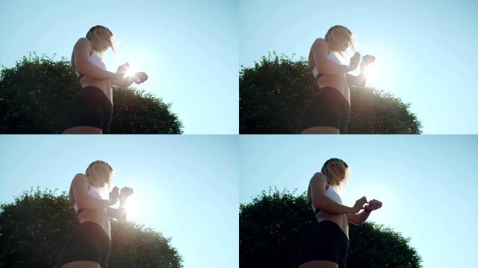 运动年轻的金发女子站在阳光下的天空背景下，检查她的智能手表的心率和距离; 户外慢跑后