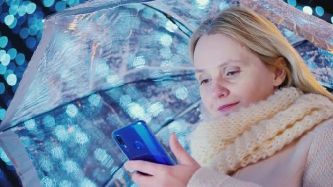 雨伞下的女人使用蓝色智能手机。在假日花环模糊的灯光的背景下