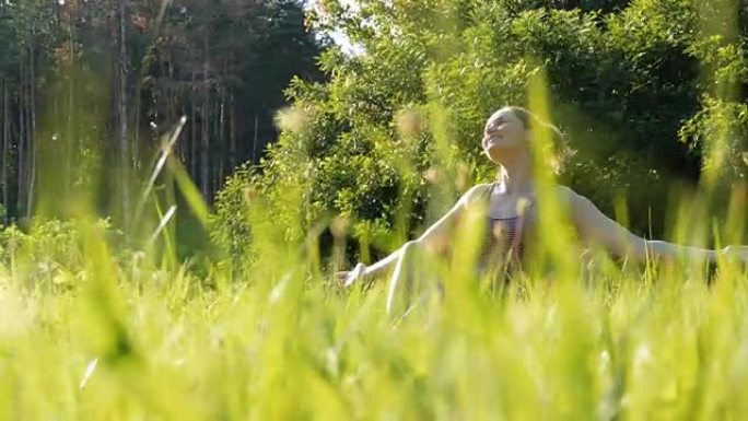 快乐的女人坐在绿色的草坪上快乐地举起双手，在日落背景下的风景秀丽的田野上