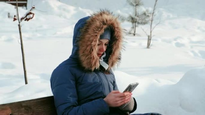 在下雪的天气里，白天，特写镜头的女人坐在长椅上，在城市的冬季公园里浏览手机。