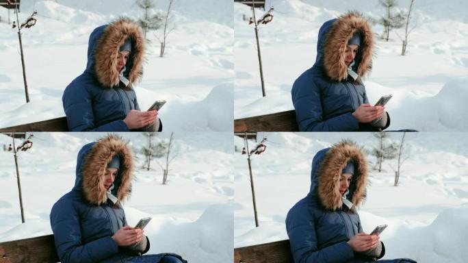 在下雪的天气里，白天，特写镜头的女人坐在长椅上，在城市的冬季公园里浏览手机。