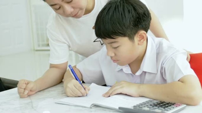 亚洲母亲的慢动作帮助儿子在白色桌子上做作业。严肃的亚洲母亲和儿子在客厅做作业。妈妈教儿子如何天才。