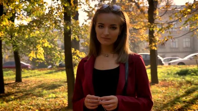迷人的年轻女性站在公园里，拿着黄叶，在美丽的晴天里若有所思地看着镜头