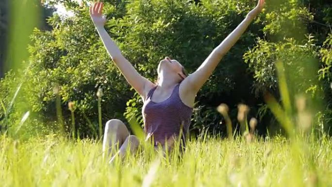 快乐的女人坐在绿色的草坪上快乐地举起双手，在日落背景下的风景秀丽的田野上