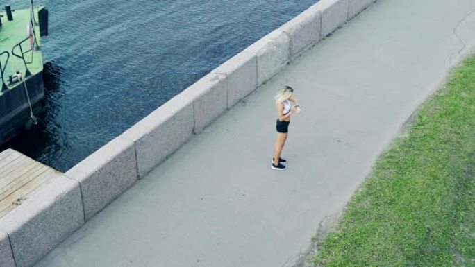 金发碧眼的女性atthlete在河边的路堤上慢动作奔跑，停下来检查她的健身追踪器的设置并进行马拉松训