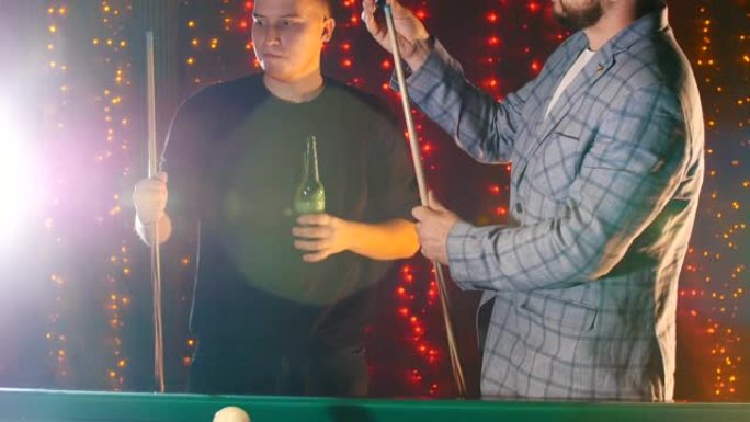 两个男人站在台球桌边，一边玩一边喝啤酒。戴眼镜的大胡子男人站在球杆上擦粉笔