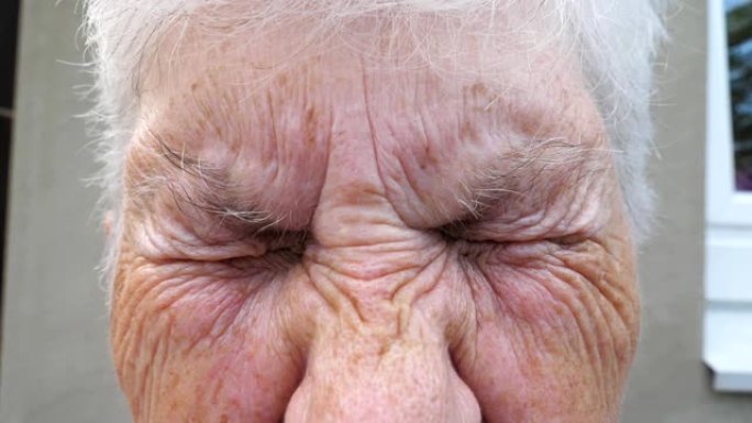 关闭老祖母皱着皱纹的脸，带着悲伤的景象看着镜头。成熟女人的肖像强烈地扭曲了她灰色的眼睛。高级女士的悲