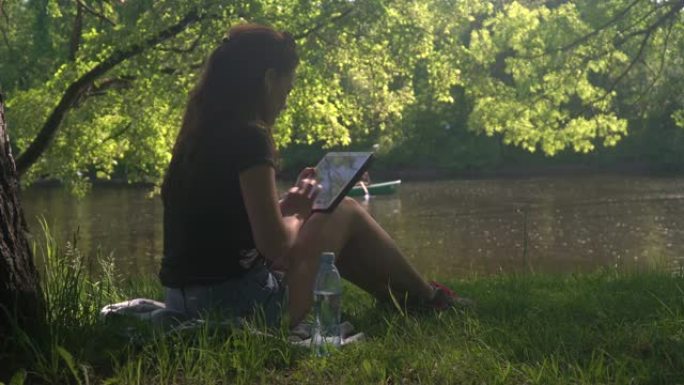 年轻女子坐在公园的河边，使用平板电脑