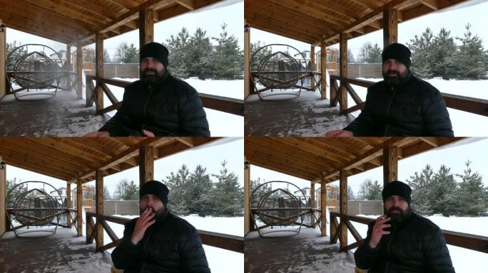 大胡子的男人抽烟，看着小屋木制露台上的智能手机。冬天松树之间的木制露台。