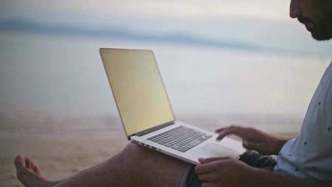 一个年轻帅气的男人在夕阳下的沙滩上用笔记本电脑工作。