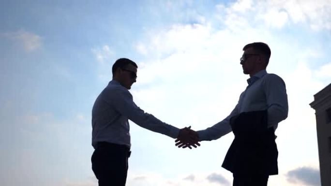 两个成功的商人在城市见面并互相问候。年轻的男同事在户外握手。户外商业伙伴握手。背景是蓝天。慢动作低角