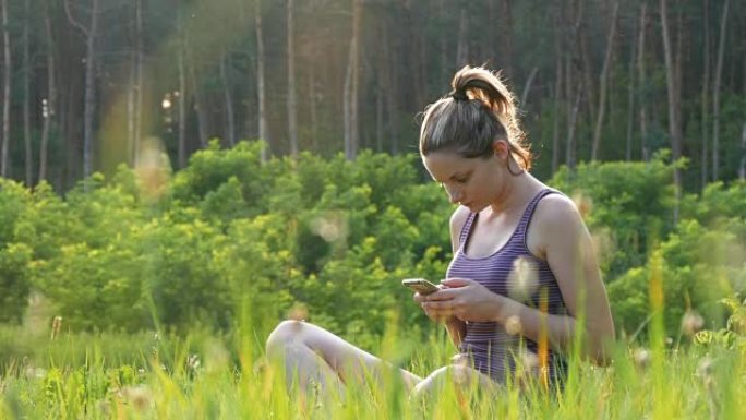 快乐的年轻女子坐在绿色的草坪上，在日落背景下在风景秀丽的田野上使用智能手机
