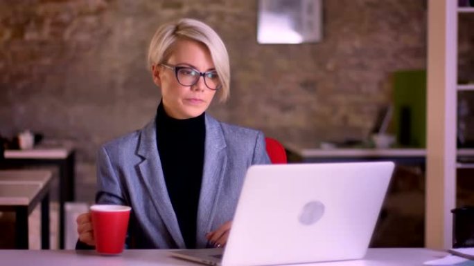 戴着眼镜的金发短发女商人的肖像在笔记本电脑的屏幕上观看并在办公室喝咖啡。