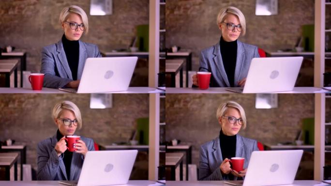 戴着眼镜的金发短发女商人的肖像在笔记本电脑的屏幕上观看并在办公室喝咖啡。