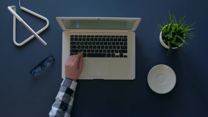 穿着灰色衬衫的创意女孩坐在桌子旁喝咖啡时正在笔记本电脑上打字。附近是点。头顶射击