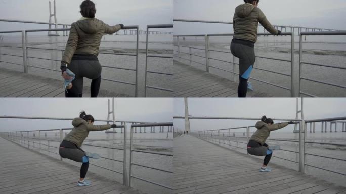 集中运动的年轻女子在带有金属栅栏的木制码头上伸展腿的慢动作镜头。