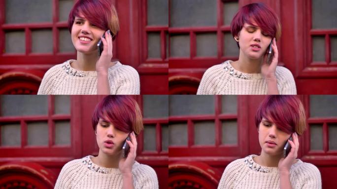 年轻的高加索粉红头发女孩的肖像在红色门背景上的智能手机上快乐地交谈。