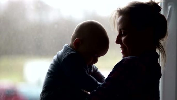 一位年轻母亲和一个六个月大的孩子在窗户背景下的剪影。产假的家庭主妇，母亲节
