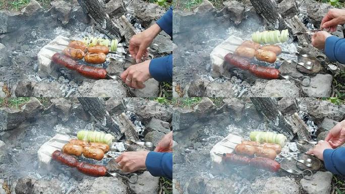 女性的手用香肠转动brochettes，特写镜头。在花园里热闷烧的余烬上烤四口香肠和洋葱。余烬在用石