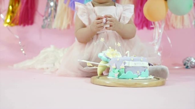 可爱的蹒跚学步的女婴吃和玩粉色柔和背景的生日蛋糕，气球和节日装饰