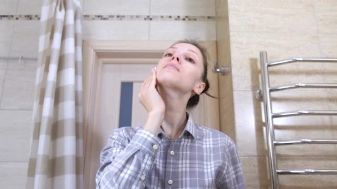 浴室里戴眼镜的昏昏欲睡的女人用化妆水用化妆棉擦拭脸。