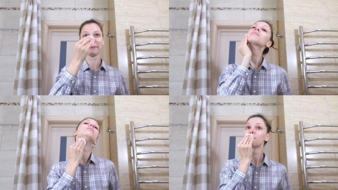 浴室里戴眼镜的昏昏欲睡的女人用化妆水用化妆棉擦拭脸。