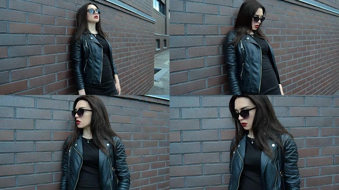 穿着黑色衣服和太阳镜的迷人年轻黑发女人站在墙上，摆一副相机