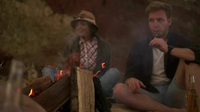 一群年轻的朋友坐在海滩上的火炉旁，喝着啤酒，烤着沼泽的棉花草。慢动作镜头