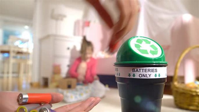 女性双手将废旧电池放入家里的专用回收箱中。孩子在后台玩耍。仅电池