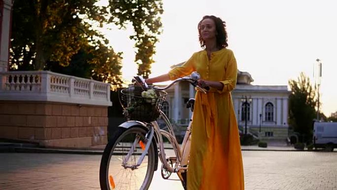 穿着黄色长裙的美丽年轻女子在黎明时拿着城市自行车的车把，篮子里放着鲜花。镜头耀斑，美丽的城市景观