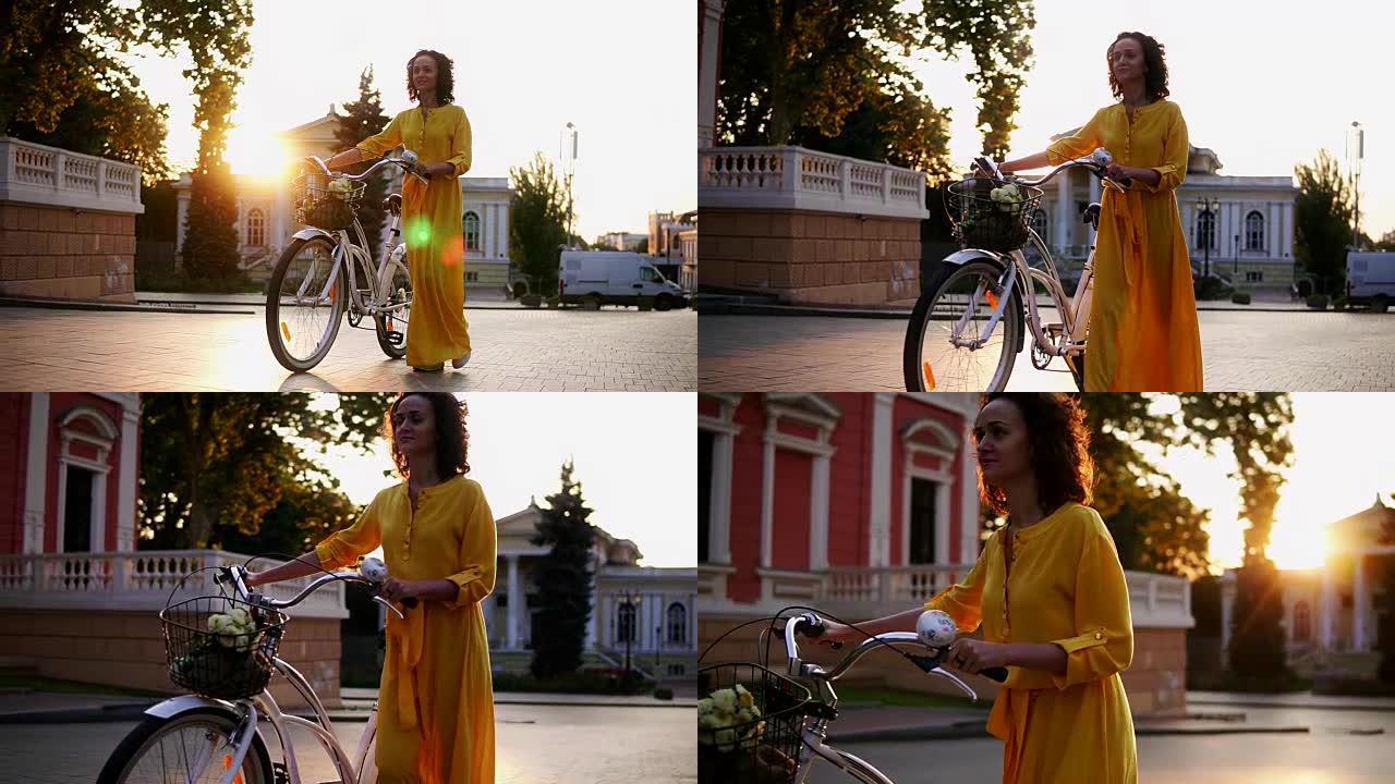 穿着黄色长裙的美丽年轻女子在黎明时拿着城市自行车的车把，篮子里放着鲜花。镜头耀斑，美丽的城市景观