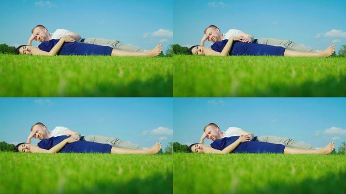 一个有怀孕妻子的男人躺在绿草地上。在蓝天的背景下