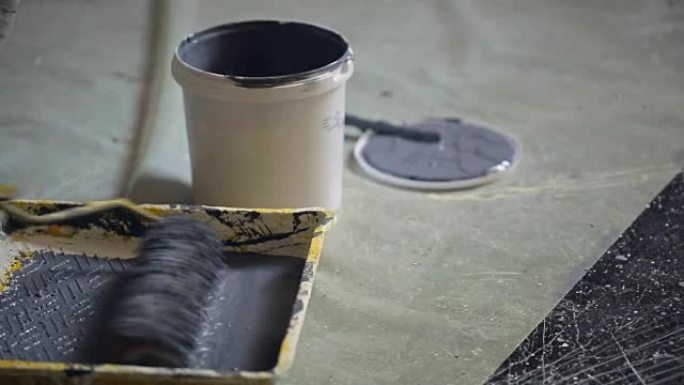 工人在罐子附近的地板上用灰色油漆将油漆滚筒浸入沟中