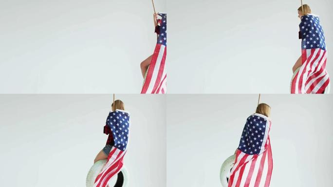 广角镜头的年轻白人女孩与挥舞美国国旗骑在一个轮胎上的摆动对白色的背景