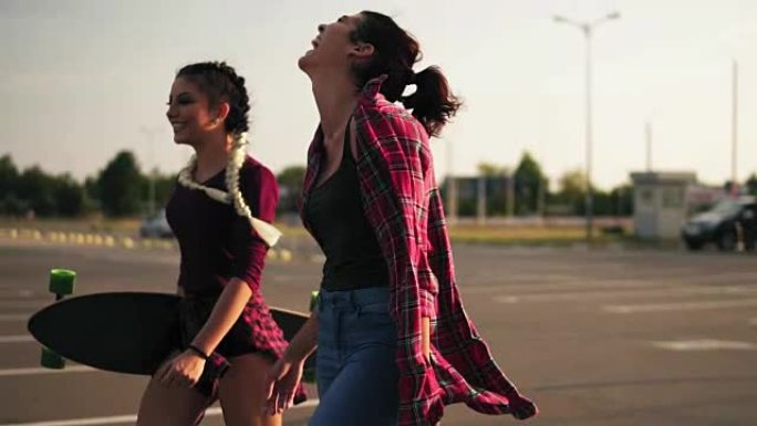 两个年轻快乐的时髦女孩在停车场一起散步，有说有笑的侧视图。一个长辫子的女孩手里拿着滑板。慢动作镜头。