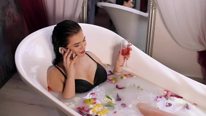 美丽的年轻女子在电话中聊天，在牛奶浴中放松身心，并从玻璃杯中喝起泡酒。4k拍摄