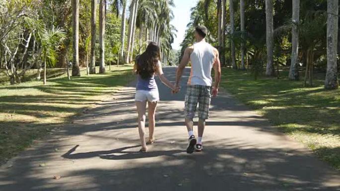 夫妻手牵着手，在热带公园散步。年轻的男人和女人沿着异国情调的公园巷，在棕榈树上观看。暑假旅行概念。后