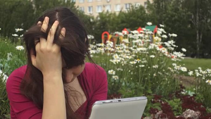 忧郁症中的年轻黑发女子在公园的雏菊旁边使用平板电脑。
