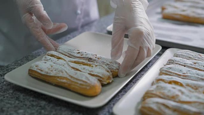 面包师用手将新鲜出炉的小饼和糖粉整齐地折叠在面包店厨房的漂亮白色盘子上。桌上有许多美味的蛋糕