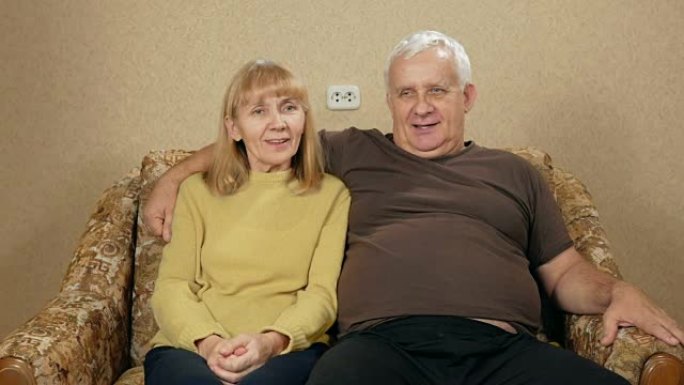 老年夫妇在家躺在沙发上休息，看着相机。他们在微笑。丈夫拥抱妻子